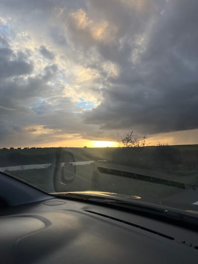 The image depicts a serene landscape viewed from inside a vehicle, likely a car, looking out through the windshield. The time of day appears to be either sunrise or sunset, commonly known as golden hour, characterized by warm, soft light.
The sky is dramatic and partially cloudy, dominating a significant portion of the scene. The low-lying sun emits an intense golden light, casting bright reflections on the vehicle's windshield and parts of the dashboard visible in the foreground. Patches of bright, golden-white clouds contrast sharply with areas of darker, grey clouds, and hints of clear blue sky are visible where the sun's rays break through.
On the horizon, silhouetted by the brilliant sunlight, is a line of trees or dense vegetation. A road or track with a visible white marking stretches into the distance, suggesting movement or travel. The dashboard of the car is visible in the lower right portion of the frame (when oriented correctly), indicating the viewpoint from inside the vehicle. The overall mood is peaceful and awe-inspiring, highlighting the natural beauty of the sky and landscape during this time of day. No specific human subjects or discernible text are visible in the image. The location context provided is Ain Johra, Morocco.