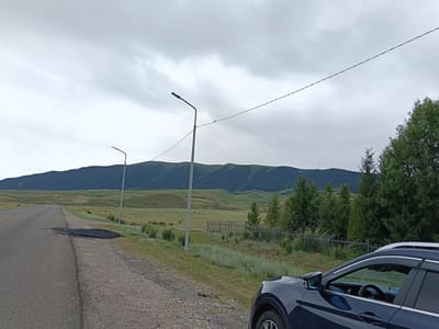 The image depicts a wide, asphalt road curving to the left, with a gravel shoulder. On the shoulder, near the road, is a patch of dark, coarse material, possibly asphalt or gravel. The landscape to the right of the road is a grassy field with scattered vegetation, leading to a fence and a stand of evergreen trees. Beyond the trees, rolling green hills and mountains form the horizon, covered in vegetation and with some evidence of human settlement in the distance. A dark blue SUV is parked on the right side of the road, partially obscuring the fence and trees. The sky is overcast with scattered clouds. The scene appears to be a roadside view in a rural or semi-rural area, consistent with the outskirts of a town like Saryozek, Kazakhstan, given the expansive natural landscape and the road infrastructure. The time of day appears to be daylight, under a cloudy sky.