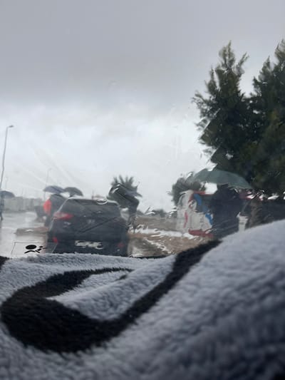 The scene depicts an exterior view through a rain-streaked vehicle windshield in Oued Jdida, Morocco, during an overcast and rainy day. In the immediate foreground, a grey and black striped fuzzy textile object occupies the lower portion of the frame, out of focus. An airbag warning decal is partially visible on the lower-left interior of the windshield.
Outside the vehicle, a wet roadway or street is visible, populated by several vehicles and individuals. A dark-colored sedan is centrally positioned, facing away from the viewer, with its rear illuminated by ambient light. Its Moroccan license plate is partially obscured by rain and reflections. Other indistinct vehicles are present in the background traffic. Several blurry human figures are discernible; at least two are using open umbrellas (one dark, one blue) while navigating or standing on the wet ground. Trees with dense, dark green foliage are visible to the right and center-right, along with a street light pole on the far left. The sky is uniformly grey and heavily overcast, indicative of current precipitation.