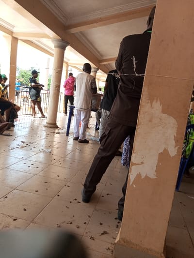 The image portrays a covered outdoor walkway or veranda with a light-colored tiled floor and structural pillars, located in Ajalli, Nigeria. The perspective is low-angle, looking through a gathering of individuals.
In the immediate foreground on the right, the lower torso and legs of a person wearing dark brown cargo pants and dark shoes are visible, partially obscured by a thick, light brown pillar. This pillar exhibits significant surface wear, with peeling material revealing irregular white patterns. A thin white string is stretched horizontally across the mid-plane of the scene, passing behind this pillar and across the individual's back.
The tiled floor in the foreground and mid-ground is covered with numerous small, dark, fragmented debris.
In the mid-ground, slightly to the left of center, a person wearing white trousers and a white patterned tunic stands with their back to the camera, positioned near a small blue stool and a dark, rectangular container, possibly a ballot box. To their left, another individual in a pink hooded sweatshirt and dark trousers also faces away from the camera, standing by a similar blue stool.
Further left in the mid-ground, a person wearing a black cap, black t-shirt, and dark shorts is standing, facing right. Several other individuals are partially visible on the far left, some seated, others standing, with some wearing yellow tops or vests.
The background shows more people gathered in a brightly lit, open area beyond the covered section, which features additional architectural columns. The overhead structure consists of a light-colored ceiling with decorative cornices. The overall scene suggests a public gathering or waiting area.