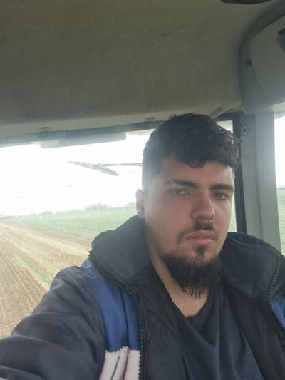 A man with dark, curly hair and a beard is inside a vehicle, possibly a tractor or other agricultural machine, given the view of a plowed field visible through the window. He's wearing a dark-colored jacket or coat with a blue interior and a darker undershirt. The interior of the vehicle is a light tan color. The location is suggested to be Lomnitsa, Bulgaria based on external information. The man appears to be taking a selfie.