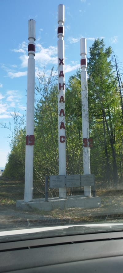 The image depicts a sign marking the location as the city of Tabaga, Russia. Three white columns stand on a concrete base. The name "ХАҤАЛАСС" is vertically written in red letters on the center column. The numbers "19" are on the left column, and "92" is on the right column, both in red. Below the columns, three informational plaques are mounted, each with text in an unknown language. Green trees are visible behind the sign, and a blue sky with scattered clouds is above. The photo appears to be taken from inside a vehicle, as part of the dashboard is visible at the bottom of the frame.