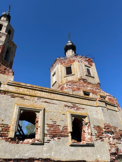 The image shows a low-angle view of a dilapidated building in Шуран, Russia, against a clear blue sky. The building appears to be a church or religious structure, judging by the presence of onion-shaped domes with crosses on top. The structure is made of brick, covered with a layer of plaster or concrete that is peeling and crumbling, revealing the brick beneath. The walls have several window openings, some of which are damaged and show signs of decay, with remnants of wooden frames visible. The overall condition of the building indicates significant neglect and disrepair.