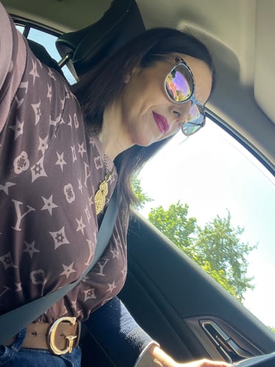 A woman is captured from the chest up, seated in the passenger seat of a car, looking downwards towards the right. She is wearing a brown long-sleeved shirt with a repeating pattern of stylized stars and flowers, possibly a designer print. A prominent gold necklace with an ornate pendant rests on her chest, and a grey seatbelt is visible across her torso. She is also wearing a tan belt with a large, gold "G" buckle. Her hair is dark and extends past her shoulders. She is wearing large, stylish sunglasses with reflective lenses, and her lips are painted with bright red lipstick. Outside the car window, green trees and a bright blue sky are visible, suggesting an outdoor setting during the daytime. The interior of the car shows a dark dashboard and door panel. The location is identified as Roodt-sur-Syre, Luxembourg.