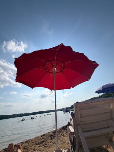 The image depicts a sunny day at a beach or lakeside in Ozyaritska-Slabadski rural council, Belarus. A vibrant red umbrella is prominently featured, angled upwards towards a clear blue sky dotted with fluffy white clouds. The umbrella's structure is visible, with its spokes radiating outwards. In the background, a white lounge chair is partially visible, suggesting a relaxation setting.
The water's edge meets a sandy shore on the right side of the frame. The calm water reflects the sky and appears to have several people in it, along with some buoys or markers. A dark green treeline borders the distant side of the water, indicating a natural landscape. The overall mood is peaceful and suggests a leisure activity, likely swimming or sunbathing, taking place on a warm day. The sun appears to be high in the sky, typical of midday. No visible text is present in the image.
