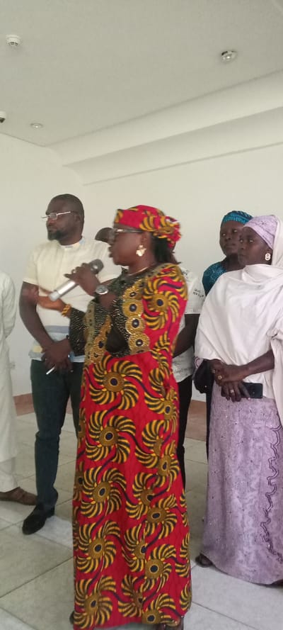 An indoor scene in Yola, Nigeria, depicts a gathering of five identifiable individuals. A woman positioned centrally, facing slightly left of the camera, is actively speaking into a silver and black microphone held in her right hand, her left hand partially raised in a gesture. She wears a vibrant red dress or gown adorned with yellow and blue spiral patterns, a matching headwrap, glasses, earrings, and a watch. The upper part of her garment features elaborate gold and black embellishments.

To her immediate left stands a man with a dark beard and glasses, wearing a cream short-sleeved shirt with a patterned collar and dark trousers. His arms are crossed, with a white object, possibly a pen or cigarette, held in his left hand. He wears a yellow beaded bracelet on his left wrist.

To the speaking woman's right, a woman in a white headscarf and a light purple, intricately embroidered gown holds a dark smartphone in her left hand, which rests upon her right hand. Rings are visible on her fingers. Partially visible between these two women are two other individuals: one with a blue patterned head covering and another largely obscured.

The environment features plain white walls and a white ceiling with recessed lighting and a smoke detector. The floor is covered with light-colored tiles.