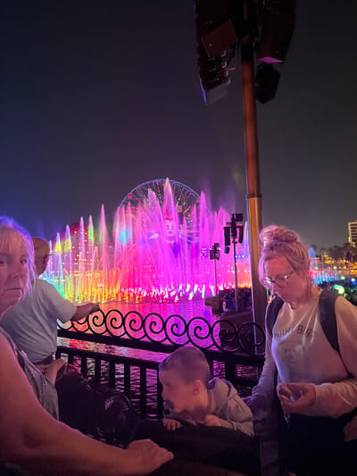 The image depicts a nighttime view of the World of Color show at Disney California Adventure Park in Anaheim, USA. Colorful fountains and lights illuminate the scene, with the Pixar Pal-A-Round ferris wheel visible in the background. A family is observing the show from behind a black metal fence. An adult with glasses and a white shirt is standing with a young child beside them, both looking towards the show. Another adult is seated with another young child behind the fence.