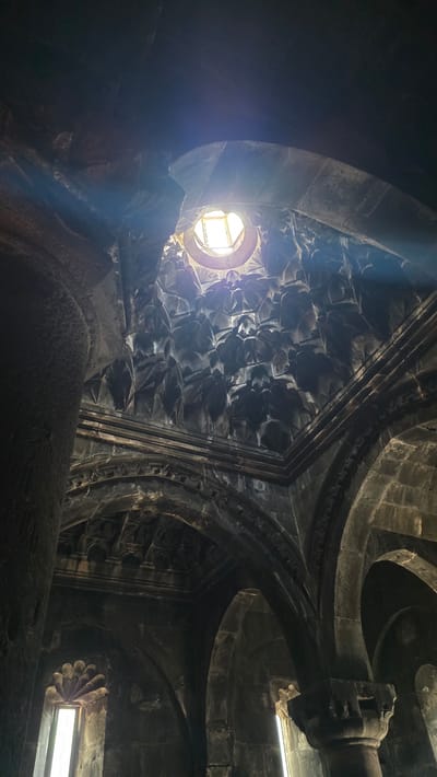 The image depicts the interior of Geghard Monastery in Goght, Armenia, focusing on the ornate stone architecture. The ceiling features a circular oculus through which sunlight streams, creating a visible beam. The dome is intricately carved with geometric patterns. Below the dome, arched structures and stone columns are visible. Two small windows with decorative stone tracery are present on the left and right walls, emitting light. The overall impression is one of ancient, well-preserved stonework within a sacred space. There are no people or animals in the image.
