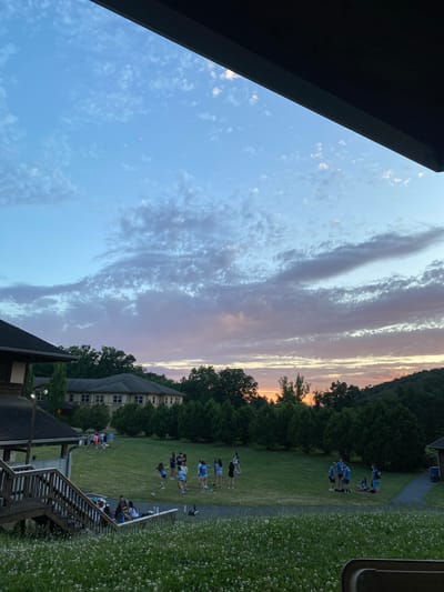 The image depicts a group of young people gathered on a grassy field in front of a large, multi-story building, likely a camp or retreat center. The scene is set during twilight, with a colorful sky filled with clouds ranging from pale blue to hues of pink and orange near the horizon. The building features wooden siding and a dark roof, with several windows visible. A wooden staircase leads down from a porch on the left side of the image. The field is dotted with small white flowers and surrounded by lush green trees. Several individuals are standing and sitting in small groups, engaged in conversation or simply relaxing. The overall atmosphere appears to be calm and social, indicative of an evening gathering. The location is identified as Washington Township, United States of America.