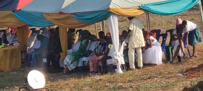 The scene depicts an outdoor gathering under a large, multi-colored canopy tent in Alayi, Nigeria. The ground consists of dry grass and exposed soil. The tent canopy features drapes of white, gold, teal, and red fabric, with white decorative fabric hanging vertically within.
Approximately 20-30 individuals, a mix of adults and children, are present. Most are seated on red, blue, and white plastic chairs arranged in rows under the tent. Attendees wear a combination of traditional Nigerian attire, including patterned dresses and head wraps, and casual Western clothing. A man in a light cream-colored traditional outfit stands upright near the center-right. Towards the far right, a young girl in a white dress is seated. Two women on the far right interact with a mobile phone; one is seated in a white dress and pink cap, while the other stands leaning over her. On the far left, a small round table draped in yellow fabric holds a floral arrangement. In the lower-left foreground, a bright, circular artificial light source on a stand is positioned on the ground, directing light towards the seated group.