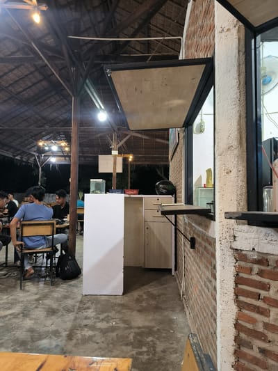 The image depicts an outdoor dining area in Cempeudak, Indonesia. Several individuals are seated at tables under a thatched roof structure supported by wooden poles. The lighting is dim, provided by overhead bulbs and decorative string lights. To the right, a brick wall features a serving window with a hinged countertop extending outwards. Behind the window, a small counter area is visible, cluttered with various items. A white cabinet unit sits adjacent to the wall, and a black motorcycle helmet rests on the counter. The floor is concrete. A partial view of a wooden table is in the foreground.