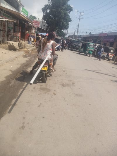 A motorcycle carries two people and a long white pipe down a sunny street. The passenger sits behind the driver, and the pipe extends far beyond the motorcycle's rear. Buildings line the left side of the street, and various vehicles, including auto rickshaws, occupy the right side. People are seen walking and standing near the vehicles, creating a busy street scene under a bright sky.
