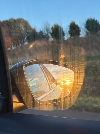 A car's side mirror reflects sunlight and what appears to be power lines or tall structures against a bright sky. The mirror's surface is dusty or dirty, obscuring the reflection slightly. The background outside the car window shows trees and blue sky, adding depth to the composition. The image is taken from inside the car, showing part of the door frame.