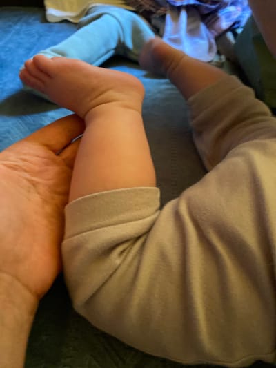 The image shows a close-up view of a baby's leg and foot, with an adult's hand holding the baby's leg. The baby is wearing light-colored pants. The background is out of focus but appears to be a bed or couch with blue and other colored fabrics. The scene is indoors. The location in the city of Korvet, Russia is not determinable from the image.
