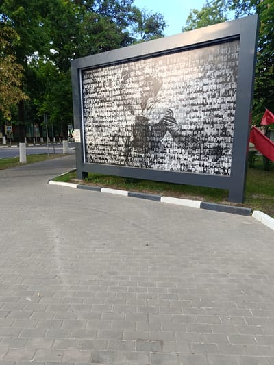 In Gubkin, Russia, a large black-framed billboard displays a black and white mosaic of numerous portraits. The mosaic is overlaid with a larger, silhouetted image of a figure holding a flag, with a portion of the mosaic forming the background. The billboard stands on a paved area composed of grey rectangular bricks. To the left of the billboard, a street is visible with a pedestrian crossing sign, white-lined curbs, and trees lining the sidewalk. Two red flags are attached to the right side of the billboard. The sky above is clear and blue.