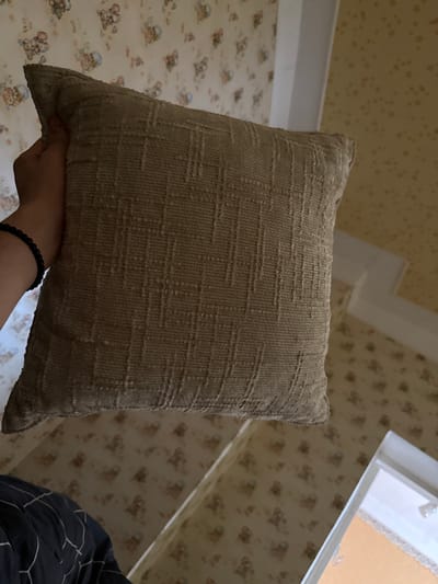 The media file shows a close-up, slightly angled view of a textured, tan throw pillow. The pillow is held by a person's hand, visible at the bottom left of the frame, and they are wearing a black beaded bracelet. The background features wallpaper with a repeating pattern of small, cartoonish bears wearing blue hats. Above the wallpaper, there is a white architectural element, possibly a ceiling molding or part of a doorway, against a textured yellow ceiling. A window frame is visible in the upper right, with what appears to be a light-colored curtain or blinds behind it. The lighting suggests it might be daytime, but the indoor setting offers no specific clues about the weather. The overall impression is of an indoor, domestic setting. No text is visible in the image.