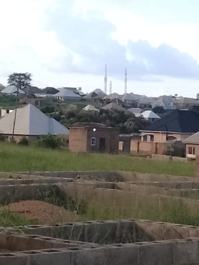 The image depicts a developing residential area in Barakin Acha, Nigeria, characterized by a mix of completed structures and ongoing construction. In the foreground, foundation outlines made of concrete blocks are visible, suggesting new homes are in the process of being built. Beyond these foundations, a gently sloping, grassy terrain is present, dotted with a few scattered trees and bushes. Several houses with diverse roofing styles, including grey and dark blue pitched roofs, are scattered across the landscape. Some houses appear to be completed, while others are in various stages of construction. A small, brown, rectangular building with a single window is centrally located in the mid-ground. In the background, more houses and buildings are visible on a slightly elevated area, with two tall radio towers piercing the overcast sky. The overall impression is one of a growing community, with a clear progression of development from foundational work to established residences.
