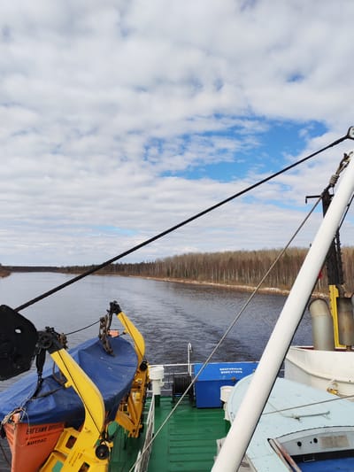 The image captures a perspective from the deck of a ship as it navigates a wide river or canal in Kurdiug, Russia. The vessel is moving along a serene waterway, flanked on both sides by dense forests of deciduous trees, currently bare of leaves, suggesting a late winter or early spring setting. The sky above is characterized by scattered white clouds against a backdrop of clear blue.
On the left side of the ship's deck, an orange lifeboat, partially covered by a blue tarp, is prominently displayed, secured within bright yellow davits. Visible text on the lifeboat identifies it as "ЕВГЕНИЙ ОРЛОВСКИЙ" (Evgeny Orlovsky) and indicates its home port or destination as "КОТЛАС" (Kotlas). Further back on the deck, a blue storage box is visible, labeled with "АВАРИЙНЫЙ ИНВЕНТАРЬ" (Emergency Inventory). A thick black cable or rope stretches diagonally across the upper part of the frame, originating from the ship's rigging on the right. Portions of the ship's superstructure, including a white mast or pole and pipes, are visible on the right. The deck itself features areas painted in green and grey. The scene depicts a calm daytime journey through a northern Russian landscape.