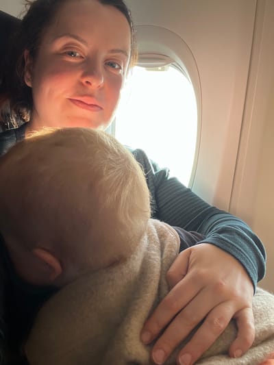 The image displays an adult female holding an infant within the interior of an aircraft cabin. The adult female, visible from the chest up, has dark hair and a neutral expression, looking directly forward. She wears a dark, striped long-sleeved garment. An infant, with blond hair, rests its head against her chest, facing away from the camera, largely obscured by a light beige blanket. The adult's left hand is visible, resting on the blanket. An oval aircraft window to the right of the subjects shows bright daylight, backlighting the scene. The specific geographical location within Babergh, United Kingdom, cannot be determined from the visual evidence provided.
