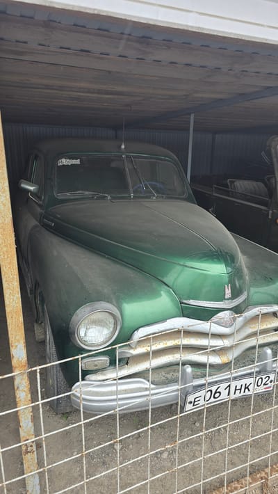 A green vintage truck is parked in a wooden structure, possibly a garage or barn. The truck is facing towards the right of the frame, with its front grill and headlights clearly visible. The grill is chrome and shows some signs of rust. A license plate with the text "E 061 HK 02" and "RUS" is attached to the front of the truck. The truck's hood is dusty, and the windshield is visible. There is a wire fence in the foreground, partially obscuring the view of the truck. On the side of the truck, near the driver's side door, a sticker with text that appears to be "НА БЕРЕГА" is visible. The setting appears to be outdoors in Bol'shoi Mashlyak, Russia, with a gravelly ground and some vegetation visible behind the fence. The lighting suggests it is daytime. No people are visible in the image.