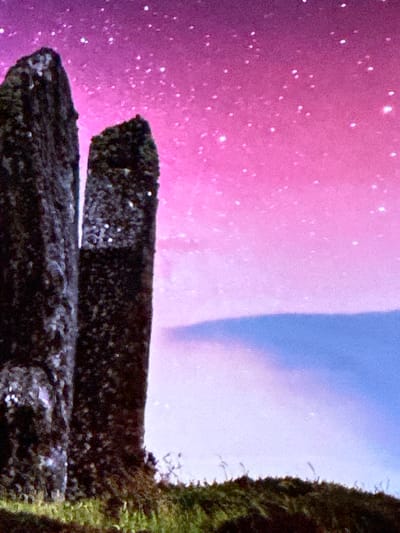 The image depicts a dramatic nighttime landscape featuring two large, dark, monolithic stones set against a vibrant, starry sky transitioning from deep purple to pink. The stones are rough-hewn and appear ancient, possibly megalithic. The sky is filled with numerous small, bright points of light representing stars, and a broad, soft band of blue and purple stretches across the middle ground, suggesting clouds or a celestial phenomenon. In the foreground, a grassy, uneven terrain is visible, with some darker shadows indicating the base of the stones and the undulation of the land. The overall impression is one of mystical grandeur and solitude. The location is identified as Villa Pasquali, Italy.