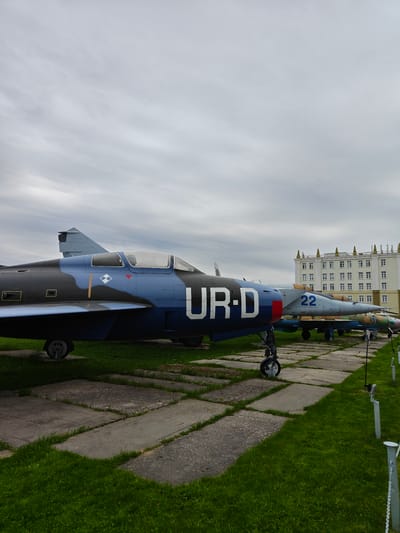 The image depicts a static display of Soviet-era fighter aircraft in Arkhangel’skoye, Russia. Dominating the foreground is an aircraft painted in a dark blue camouflage scheme with the large white letters "UR-D" emblazoned on its fuselage. Adjacent to it is another aircraft painted in a light blue/gray color with "22" on the side. Both aircraft are situated on concrete slabs interspersed with grass. In the background, a large, multi-story building with numerous windows can be seen under a cloudy sky. There is no visible presence of people. The scene is presumably within a museum or memorial dedicated to military aviation. A metal barrier and chain is present to the right of the image.