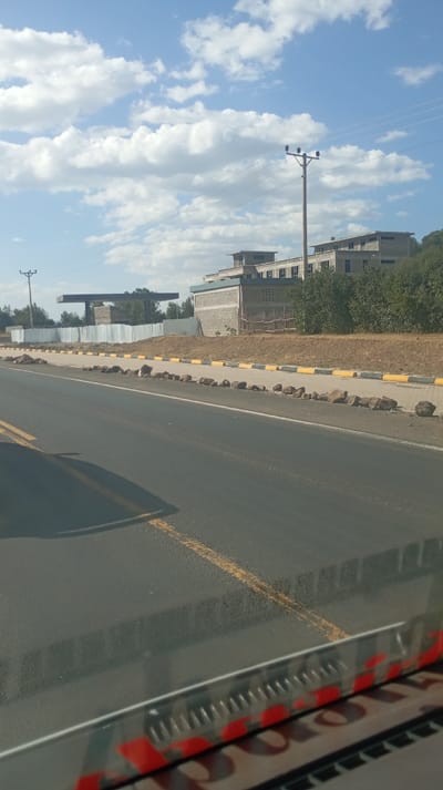 A road stretches into the distance beneath a partly cloudy sky. Along the roadside, a row of large rocks borders a paved walkway marked with yellow and white stripes. In the background, structures include a gas station canopy, a gray building with multiple levels, and utility poles with visible wires. The view is likely from inside a vehicle, as part of the dashboard is visible at the bottom of the frame.