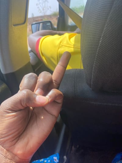The image captures an interior view from a vehicle in Unguwar Gaya, Nigeria, under bright daylight conditions. In the foreground, a dark-skinned left hand is visible, positioned with its palm facing upward and slightly towards the viewer, with the middle finger extended in a pronounced gesture. A small, bright point of light, consistent with a sun reflection, is evident on the palm. To the right, a black fabric car seat headrest occupies a significant portion of the frame. Beyond the hand and headrest, the upper body of an individual wearing a bright yellow short-sleeved shirt with red trim is partially visible, seated in a forward position. Through the vehicle's open window and side mirror, a hazy outdoor environment is discernible, featuring distant green vegetation and a light-colored, possibly dirt or sand, landscape under a bright sky. A small section of blue fabric patterned with white and red flowers is visible at the bottom left edge of the frame.