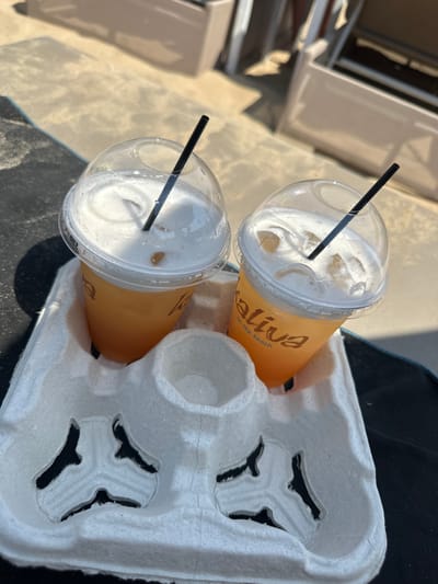 Two clear plastic cups with dome lids and black straws are filled with an orange beverage and topped with foam. The cups are placed in a white paper beverage carrier. The cups are labeled with "Maliva on the beach." The carrier and cups are positioned on a dark surface, possibly a towel, with a sandy or concrete-like background. In the blurred background, light-colored lounge chairs are visible. The lighting suggests an outdoor, daytime setting, likely a beach or poolside area in Ayia Napa, Cyprus.