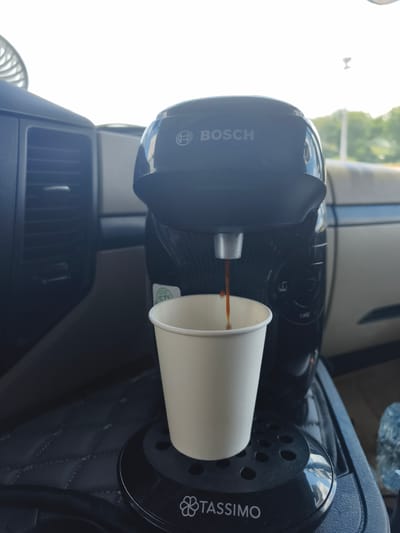 In Ciry-le-Noble, France, a Bosch Tassimo coffee machine is actively brewing coffee into a white paper cup. The coffee machine, primarily black, is positioned on a quilted dark gray surface, likely a car's interior console or seat. The machine displays the "BOSCH" logo and the "TASSIMO" brand name on its base. To the left of the coffee machine, a dark gray air vent is visible, indicating the setting is likely within a vehicle cabin. The background shows the interior of a vehicle with beige upholstery and a glimpse of greenery and sky through a window. The scene captures the process of freshly brewed coffee dispensing into the cup.
