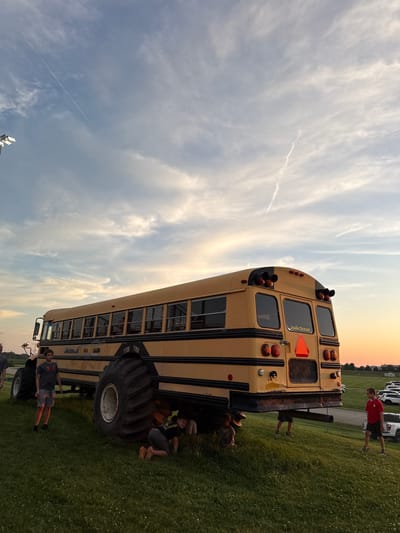 The image shows a modified yellow school bus with oversized tires, parked on a grassy incline. The bus has "JOHN DEERE" written on the back. Several people, appearing to be children and teenagers, are visible around the bus. One person in a red shirt and black shorts stands to the right, looking towards the bus. Another person in a grey t-shirt and grey shorts stands to the left, also looking at the bus. Several other figures are partially obscured or near the ground under the bus. The sky above is a mix of blue with wispy clouds and hints of sunset colors. The environment appears to be an open, grassy area, possibly a field or park, with a parking lot visible in the background to the right. The image does not contain any information that would place it in Joliet, United States of America.