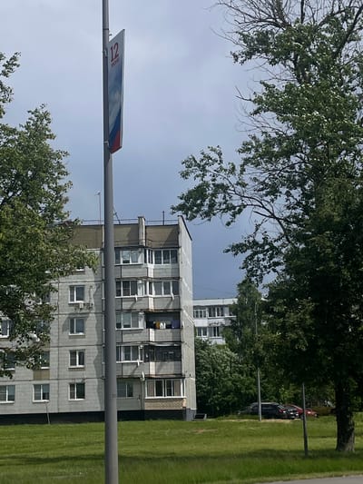 The image depicts a residential scene in Yakovlevskoye, Russia. The primary subjects are buildings and trees, suggesting an urban or suburban environment. A tall, multi-story apartment building, characterized by its gray, tiled facade and numerous windows, dominates the central portion of the frame. Balconies are visible on some floors. To the right of the building, a well-maintained, vibrant green lawn stretches out, indicating it is likely daytime and fair weather, possibly summer or late spring due to the lushness.
In the foreground and framing the scene are mature trees with dense green foliage, suggesting a warm season. A street sign, which appears to be a bus route marker, is attached to a pole crossing the image horizontally. The sign is rectangular and white, with blue and red stripes along its border. Text on the sign is visible but partially obscured, though the numeral "12" is clearly legible.
Behind the apartment building, more trees and other structures are visible, including another apartment building further in the distance. Several cars are parked on the grass or along the edge of a road, partially obscured by trees and foliage, indicating a residential area where parking is available. The overall impression is one of everyday life in a Russian residential district. The lighting suggests it is daytime, with a bright sky and natural light illuminating the scene. No people are directly visible, and the scene is peaceful and static.