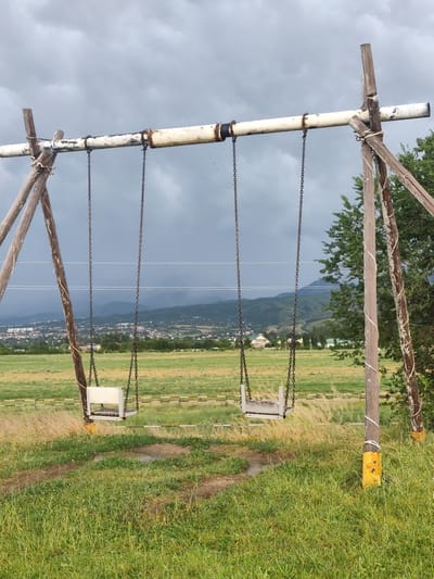 This is a full shot of a rustic swing set in an outdoor, grassy environment. The swing set is constructed from weathered wooden posts and a metal pipe crossbar, with two chains hanging down, each supporting a white swing seat. One swing is to the left, and the other is to the right, both appearing unoccupied. The scene is set against a backdrop of rolling hills and a town, identifiable as Dolan, Kazakhstan, with a cloudy, overcast sky. The foreground is dominated by lush green grass, with some areas appearing damp or muddy. The overall atmosphere is somewhat melancholic and still. There are no people or animals visible in the image.