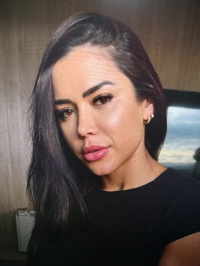 This is a close-up, headshot of a woman with dark, straight hair that falls over her shoulders. She is looking towards the camera with a slight smile. Her makeup includes bold eyebrows, dramatic eyelashes, and pink lipstick. She is wearing a simple black top and a small gold hoop earring with a clear stone in her left ear.
The background is out of focus, but appears to be an indoor setting with a textured wall on the left and a darker, possibly reflective surface on the right. The lighting suggests it could be daytime, but it is difficult to determine the exact time of day or weather conditions. There is no visible text in the image. The woman's expression is pleasant and confident, and the overall impression is of a casual, personal photograph.