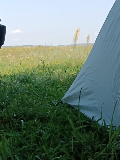 The image depicts a close-up view of a light blue tent pitched in a grassy field. The foreground is dominated by lush green grass, with some blades appearing in sharp focus. In the middle ground, the tent occupies the right side of the frame, its fabric creased and textured. Beyond the tent, a field of dry grass and wildflowers stretches towards the horizon. Two tall, feathery stalks of grass stand out in the mid-distance. The sky is a pale blue with a few wispy clouds. On the far left edge of the image, a dark, indistinct object, possibly part of a vehicle, is visible. The scene suggests an outdoor setting, likely a campsite in a rural or natural environment. There is no indication of the specific location within the city of Uran, Russia.