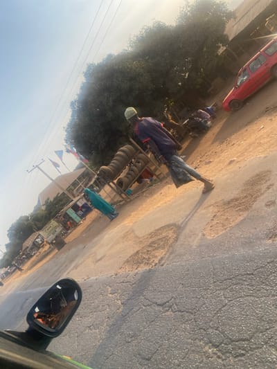 A roadside scene shows a man standing by a cart filled with stacked tires. He wears a blue shirt and a green cap. Shops line the street, which is partially paved and partially dirt. A red car is visible in the distance. The image is taken from inside a vehicle, with a side mirror visible in the foreground, reflecting the photographer. The scene is bathed in sunlight.