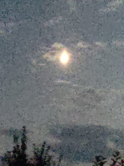 The image shows a blurry, pixelated sky with a bright, circular object in the upper center, possibly the sun partially obscured by clouds. There are dark, indistinct shapes of trees or foliage at the bottom of the frame, suggesting an outdoor scene. The overall impression is of a photograph taken with low resolution or in poor lighting conditions. The location of Арчалы, Kyrgyzstan, is mentioned, but the image itself does not provide any specific visual cues to confirm or deny this location.