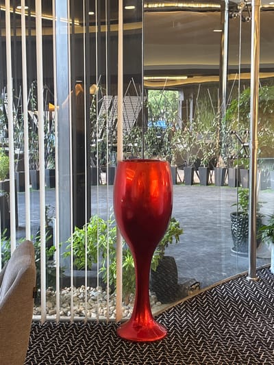 In the city of Khuraybah Al-Souk, Jordan, a large, bright red glass vase, shaped like a wine goblet, stands prominently on a patterned black and white carpet. The vase is positioned in the foreground, to the right of a grey armchair. Behind the vase, a glass partition reveals an outdoor area with numerous potted plants. The plants are in dark grey, square planters arranged in rows. The ground outside appears to be paved or tiled. Vertical white blinds are visible through the glass, obscuring parts of the outdoor view. Reflections on the glass show interior lighting and architectural elements. The scene suggests an indoor seating area adjacent to a garden or patio, possibly in a restaurant, hotel, or commercial establishment.