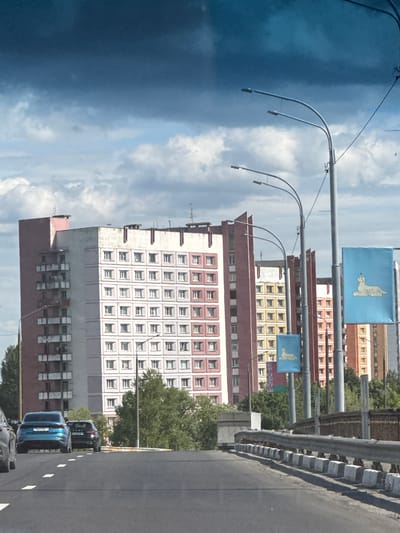 The image captures a view from a vehicle on a road, likely a bridge, looking towards a row of tall, multi-story apartment buildings. The architecture of the buildings suggests a Soviet-era style, characterized by repetitive window patterns and balconies. One building is predominantly pink and white, while others are a mix of pink, red, and yellow. Green trees are visible in front of and between the buildings. Several streetlights with curved poles are present, and two blue flags with a stylized depiction of a white animal, possibly a lynx, are hanging from poles. The sky is partly cloudy with dramatic, dark clouds interspersed with lighter areas. In the foreground, the road has white dashed lines and a metal guardrail. Several cars are visible on the road, including a blue car in the left lane and a black car behind it. The scene appears to be a typical urban or suburban landscape within the Красненскі сельскі Савет, Belarus.