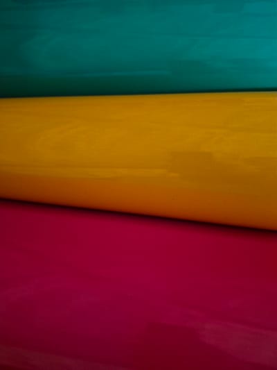 The image shows a close-up, horizontal view of three rolled-up pieces of fabric, stacked on top of each other. The top fabric is a deep teal color, the middle fabric is a bright yellow-orange, and the bottom fabric is a vibrant magenta. The rolls are positioned diagonally from the upper left to the lower right of the frame. The lighting creates subtle shadows and highlights, suggesting a slightly textured surface on the fabrics. The background is dark and out of focus. This image does not depict any specific location in Masów, Poland.