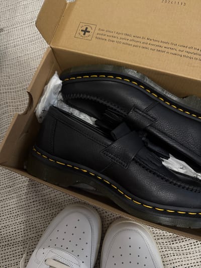 The image displays an indoor scene, likely in a home in Hainburg an der Donau, Austria, featuring a pair of new black Dr. Martens tassel loafers resting inside their open cardboard shoebox. The loafers are made of pebbled leather, characterized by their prominent yellow stitching around the black sole and decorative tassels on the vamp. White tissue paper is visible within the box, cushioning the shoes.

To the right of the shoebox, the front and side of a white sneaker, likely a Nike Air Force 1 based on its design with a perforated toe box and white laces, are partially visible. The objects are placed on a light-colored, textured rug or carpet.

The inside flap of the brown cardboard box features visible text and branding. A date or product code "20241113" is printed, along with the circular "DR. MARTENS" logo. A historical text about the brand is partially legible: "Ever since 1 April 1960, when Dr. Martens boots first rolled off the p[roduction line, they gained a reputation among] postal workers, police officers and everyday workers, our reputation [became] folklore. Over 100 million pairs later, our belief in making things to la[st remains]." The scene suggests an unboxing or casual display of new footwear.