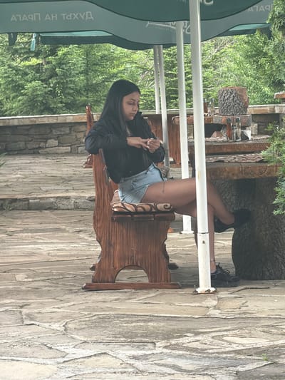 An outdoor scene in Sushina, Bulgaria, shows a woman sitting on a rustic wooden bench or chair under the shade of a large green umbrella. The woman, with long dark hair, is dressed in a dark long-sleeved top and light denim shorts, with black sneakers on her feet. Her legs are crossed, and she appears relaxed, with her hands clasped in front of her. Rings are visible on her fingers, and a small, dark tattoo can be seen on her upper left thigh.

The setting suggests a cafe or restaurant patio. Behind the woman, a sturdy wooden table or structure, possibly made from a tree trunk, holds several glass bottles, likely containing spirits or condiments. The background is lush with green foliage, indicating a natural, perhaps garden-like, environment. The ground and surrounding structures consist of irregular stone paving and textured stone walls, contributing to a natural, rustic aesthetic.

Visible on the green umbrella, in Cyrillic script, is the text "ПАЛАЦ НА ДУХА," which translates to "Palace of the Spirit." The time of day appears to be daytime, with ample natural light suggesting clear weather. The overall mood is calm and serene.