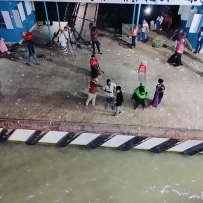 The image captures a bustling scene at what appears to be a pier or ferry terminal, likely at night or dusk, given the artificial lighting. Numerous people, both adults and children, are present on a concrete surface that shows signs of wear and scattered litter.
On the far left, a blue and white building or structure with rectangular panels is visible. Along the edge of the pier, black and white striped markers indicate the water's edge, where murky, greenish-brown water flows. Black rubber fenders are positioned along the dock, suggesting a docking area for boats or ferries.
In the left foreground, a person walks carrying a red basin on their head, indicating they might be a vendor or transporting goods. Several individuals are scattered across the scene; some stand in groups, appearing to converse or wait. A man in a maroon shirt stands near the center, while another man in a red shirt sits on a low object, possibly a stool. Further to the right, a person in a bright green outfit and another in a purple patterned dress are visible. In the background, other figures move or stand, some near a ramp-like structure. A large, round, green object, possibly a bundled item or produce, rests on the ground in the mid-background. A small red stool is also visible on the right. The overall impression is one of movement and gathering at a busy transport hub. No discernible text is visible.