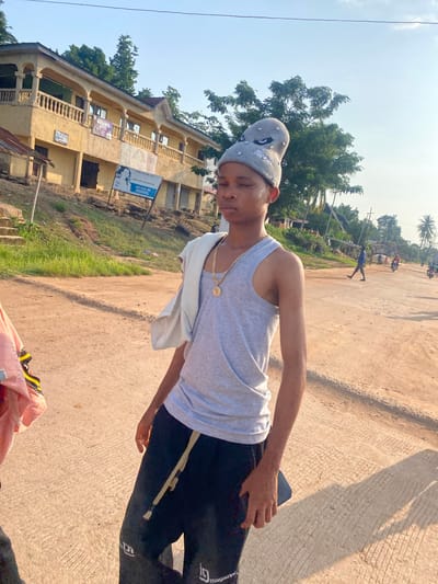 The image depicts a rural or semi-urban street scene in Nguzu Edda, Nigeria, under bright daylight conditions. In the foreground, slightly left of center, stands a young male. He has light skin, dark hair (mostly covered), and is wearing a light gray knit beanie adorned with a stylized, angry-looking character design featuring black eyes, a white eyebrow, and studded accents. He wears a light gray tank top and black trousers with a light-colored drawstring or belt hanging from the waist. A prominent gold chain with a circular gold pendant rests on his chest. His eyes are closed or heavily squinted, and his head is slightly tilted. His left hand, with a dark-colored object resembling a phone or wallet, rests against his leg. A white garment is draped over his right shoulder.
In the midground and background, an unpaved, light-brown dirt road extends into the distance, showing some faint tire track patterns. To the left, a grassy hillside with steps leads up to a two-story building. This building features yellow-painted walls, an elaborate balcony with balustrades on the upper level, and multiple windows. A sign partially obscured but reading "DUNAMIS" is visible on the upper floor balcony. Another large blue and white sign, possibly an advertisement or political poster, is positioned on the ground in front of the building. Further down the road, several individuals, mostly adult males, are visible. One man in a blue shirt walks towards the left, while others on motorcycles are further in the distance. Palm trees and other green foliage are scattered throughout the background under a clear, light blue sky with minimal cloud cover. The overall impression is a sunny, warm day in a developing community.
