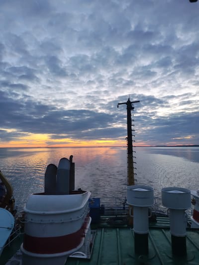 The image captures a serene view from the stern of a ship sailing on a calm body of water, likely at sunrise or sunset, given the warm light on the horizon. The sky is mostly covered with dense, textured clouds, creating a dramatic backdrop. Towards the distant horizon, a vibrant band of orange and yellow light stretches across the sky, reflecting beautifully on the water's surface.
In the foreground and midground, various parts of the ship are visible. On the left, a large structure with white and red banding, possibly part of a funnel or superstructure, can be seen, with two grey exhaust stacks rising behind it. On the right, a tall, dark mast, equipped with numerous rungs or steps and horizontal crossbars, stands prominently against the colorful sky. A light fixture or sensor is attached near the top of the mast. The ship's movement is indicated by the distinct wake trailing behind it in the water.
On the green-colored deck in the lower right, two white, cylindrical vent-like structures are present. One of these structures clearly displays the Russian text "АККУМУЛЯТОРНАЯ", which translates to "Battery Room" or "Accumulator." The scene is devoid of people, emphasizing the vastness of the water and sky. The tranquil setting and soft, low light evoke a peaceful atmosphere near Gorushka, Russia.