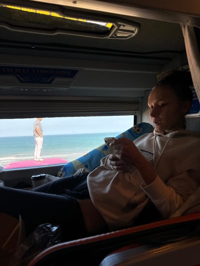 This media file shows two people inside what appears to be a bus or van, with a view of the ocean and beach through the windows. The image is oriented vertically, suggesting it was taken with a phone held sideways.

The primary subject in the foreground, a person wearing a light-colored hooded sweatshirt and dark pants, is partially visible and is looking down, holding a small object, possibly a phone or remote. Their face is illuminated from above, casting shadows. They are seated on a blue cushion with a cartoon character on it.

In the background, seen through the window, a second person is standing on what looks like a pink mat on a beach. This person is wearing a patterned short-sleeved shirt and light-colored pants. The ocean is visible behind them, and the sky is bright, suggesting daytime, possibly late morning or early afternoon given the light.

Visible text on the side of the vehicle interior reads "AM HAI LIMOUSINE". The interior of the vehicle is dimly lit, with some yellow lighting visible on the left side. The overall impression is of a casual journey, possibly a transfer to a beach destination. The lighting and the presence of the ocean and beach suggest a tropical or sub-tropical climate. The image conveys a relaxed atmosphere, with one person engaged with a device and another enjoying the view.