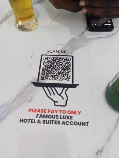 The image shows a white table top with a sticker placed on it. The sticker features a black and white drawing of a hand holding a tray with a QR code on it. The text on the sticker reads "SCAN ME" above the QR code and "PLEASE PAY TO ONLY FAMOUS LUXE HOTEL & SUITES ACCOUNT" below. There is a partially visible glass of beer to the left of the sticker and a partially visible green bottle to the right. The image was taken in Ujevwu, Nigeria, at the coordinates 5.472 latitude and 5.817 longitude. A black smartphone is visible in the upper right corner of the image.