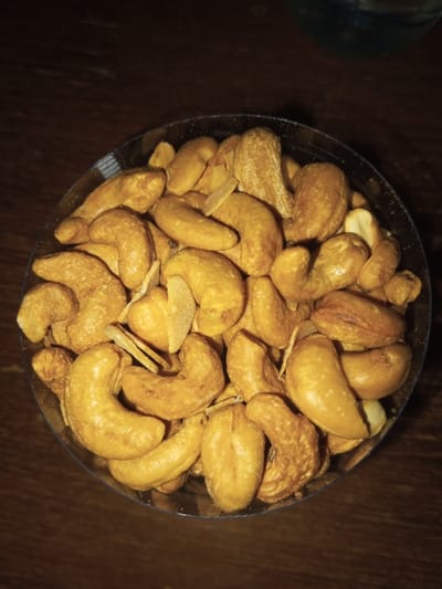 A top-down view showcases a circular container filled with cashews. The nuts are roasted to a golden-brown color, and their curved shapes are prominent. Scattered throughout the container are a few thin, crispy chips. The container rests on a dark, wooden surface, suggesting a table or countertop. Part of a bottle or container is visible in the upper right. The lighting in the scene is subdued.