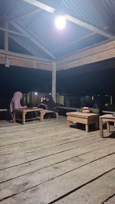 The image depicts a dimly lit outdoor wooden platform with three individuals seated at small wooden tables. A bright light bulb hangs from the corrugated metal ceiling, illuminating the scene. In the foreground, the wooden planks of the platform are visible. The individuals are all wearing hijabs and appear to be engaged in reading or studying, with books and papers on their tables. The environment suggests a rustic or simple setting, possibly a communal gathering space or outdoor classroom. The location is identified as Grong-grong, Indonesia.