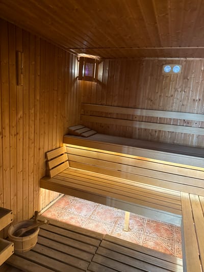 The image shows the interior of a sauna, possibly in Tulovo, Bulgaria, given the prompt's context. The room is entirely clad in light-colored wood planks, forming the walls, ceiling, and floor. A vent is visible on one wall. A small wooden bucket with a ladle sits on the floor. The room appears to be angled, potentially due to the camera's position or the design of the sauna itself. A doorway reveals a room with patterned tiling.